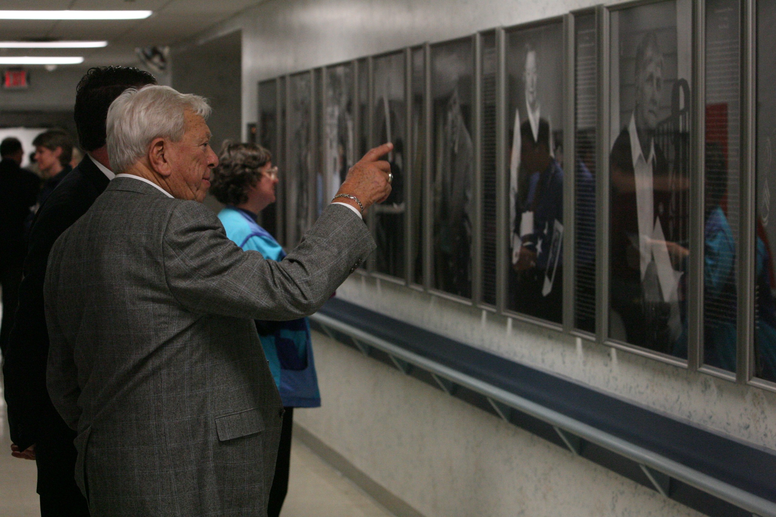 A veteran pointing toward the long installed wall of Michigan Veterans of Five Wars portrait-and-text panels at the Grand Rapids Home for Veterans