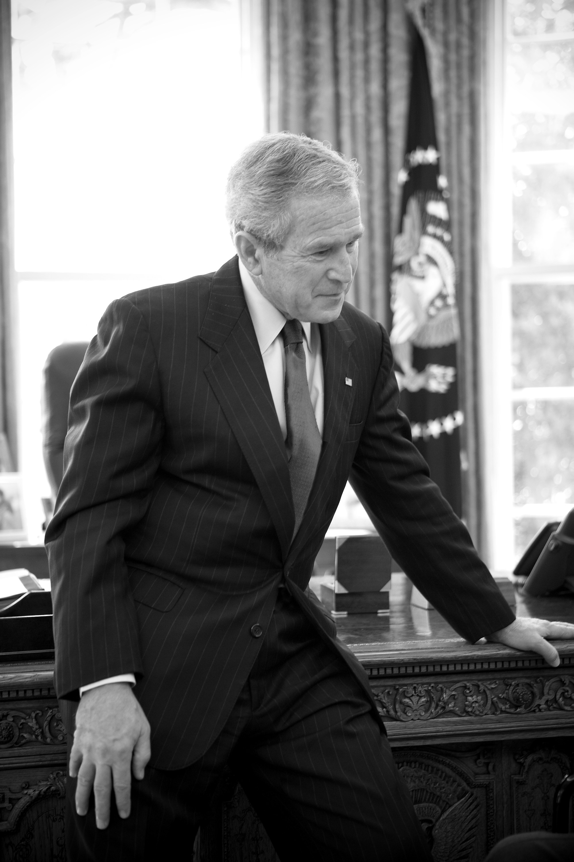 Black-and-white portrait of President George W. Bush leaning against the Resolute desk in the Oval Office