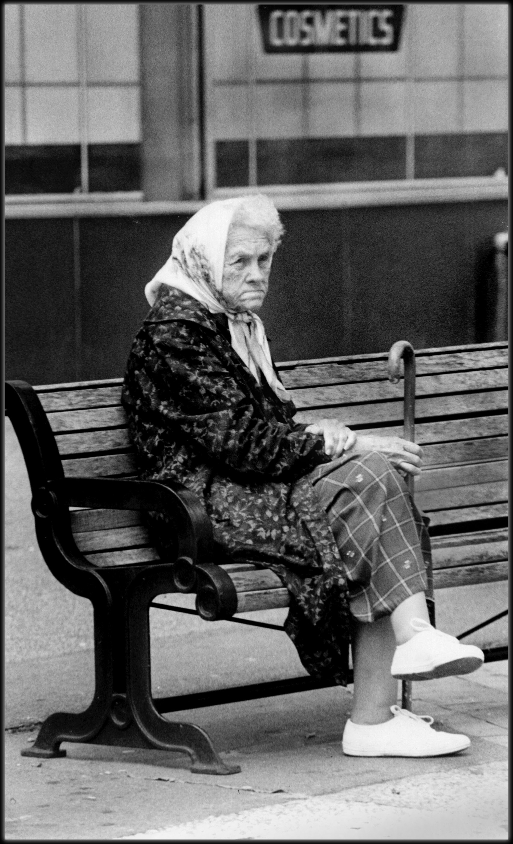 An older woman seated beneath a storefront sign reading Cosmetics