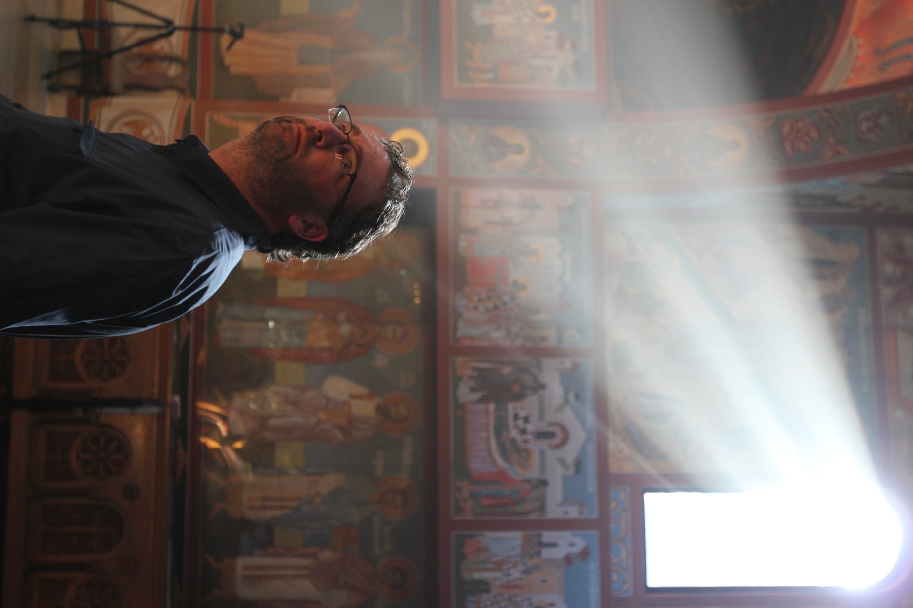 David DeJonge in the left third of the frame, looking upward beneath a shaft of window light inside New Gračanica Serbian Orthodox Monastery